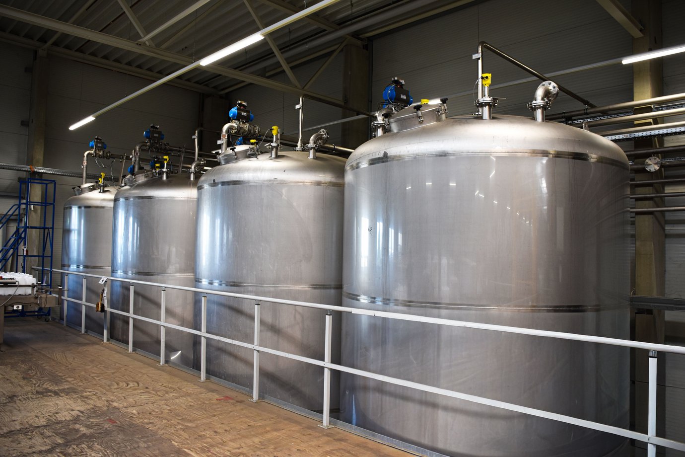 Vier große metallfarbene Tanks in einer Halle mit Brüstung im Vordergrund.